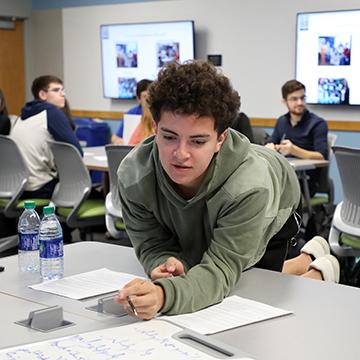 A student leans across a work table to see what another student has written.